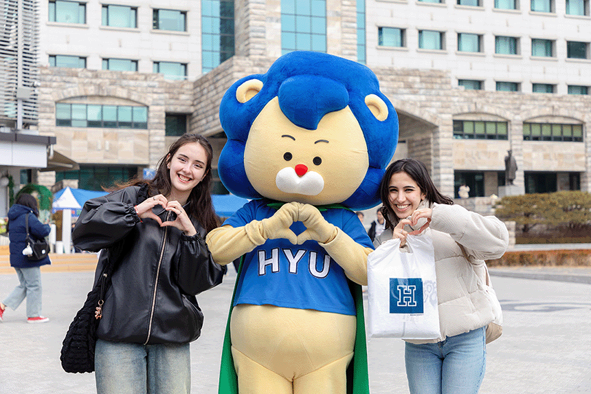Two smiling students and a blue lion mascot posing with heart hand gestures at Hanyang University in Seoul, South Korea.