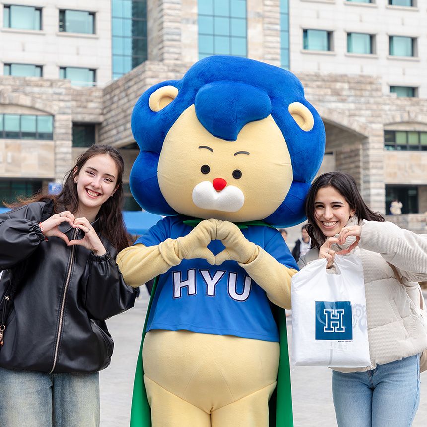 Two smiling students and a blue lion mascot posing with heart hand gestures at Hanyang University in Seoul, South Korea.