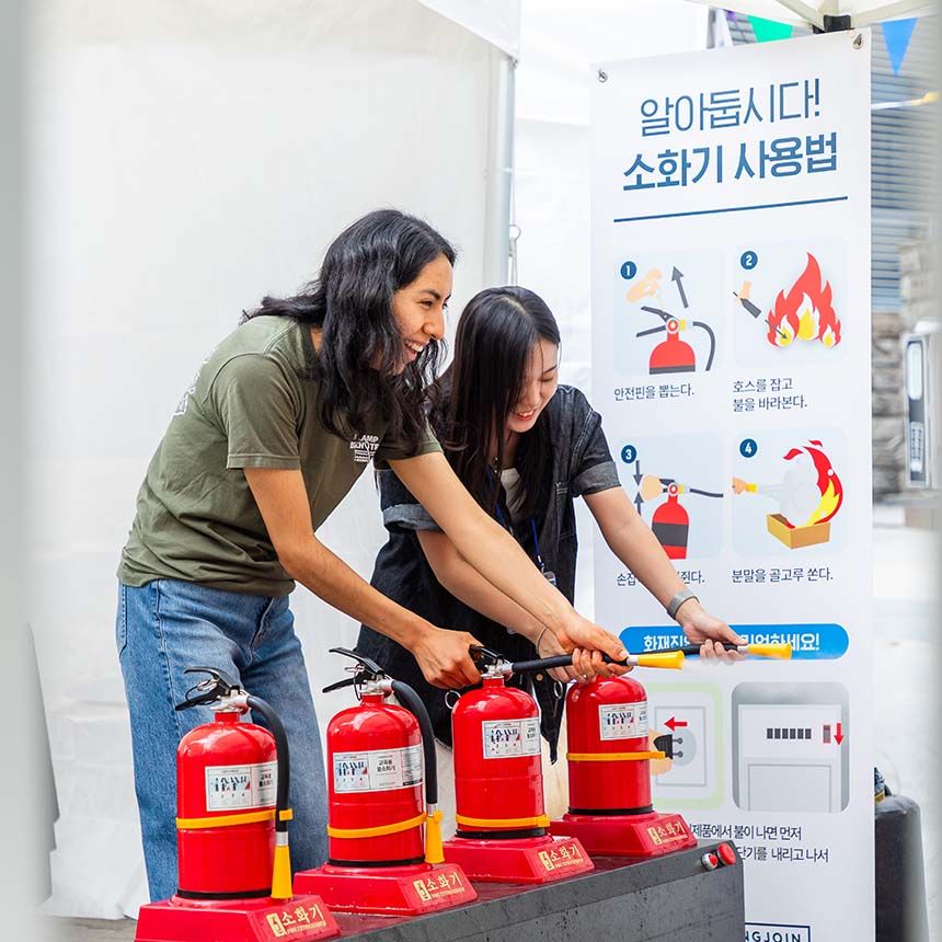 Two students smile while practicing using fire extinguishers at an outdoor safety demonstration booth with instructional posters.