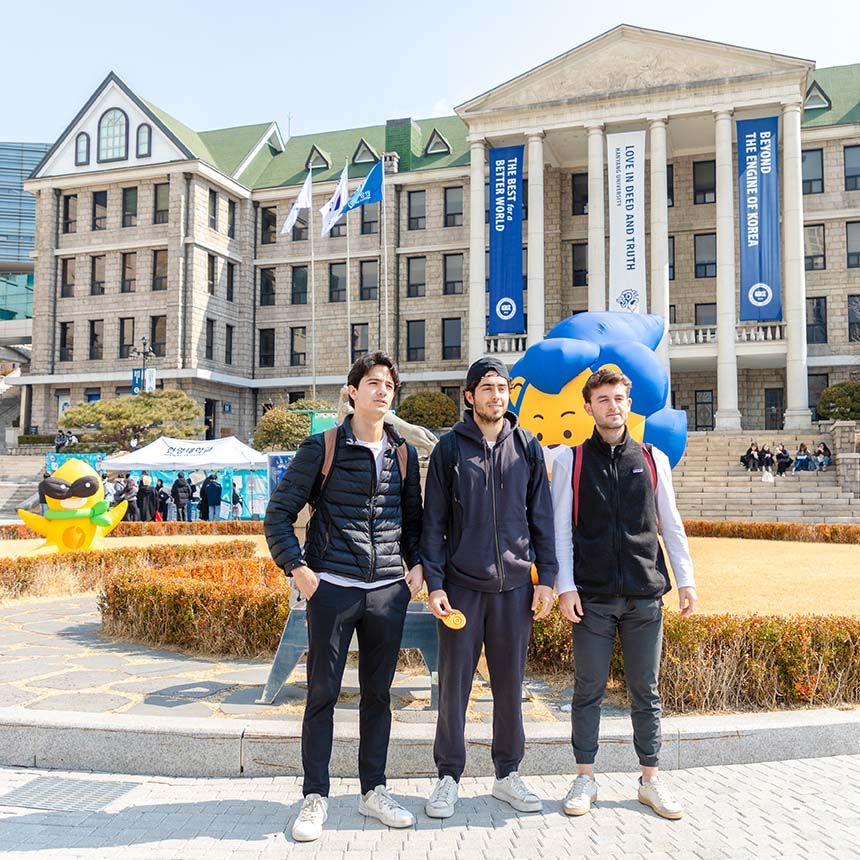 Three students posing in front of a grand stone university building with large blue banners and an inflatable lion mascot.