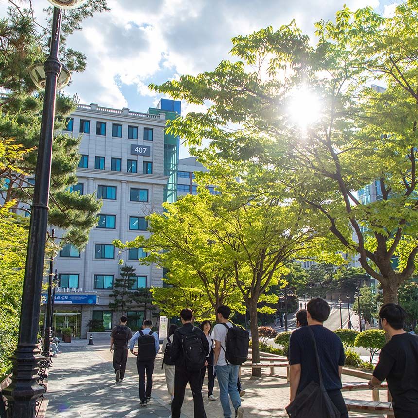 Sunlight filters through green trees as students walk along a paved campus pathway toward Building 407 at Hanyang University.
