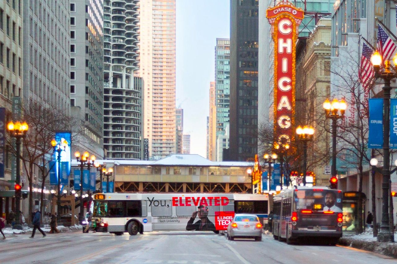 A CTA bus displaying an ad from Illinois Tech's You, Elevated campaign crosses State Street on Lake Street near the Chicago Theater.