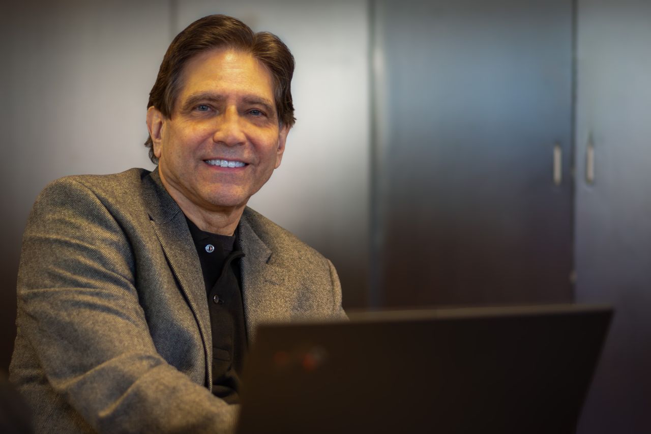 A man in a tweed jacket and dark shirt smiles in a portrait. 