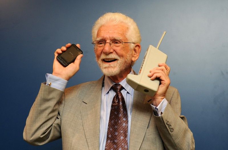A man holds a large early cellphone in his left hand and a smaller, more modern cellphone in his right hand.