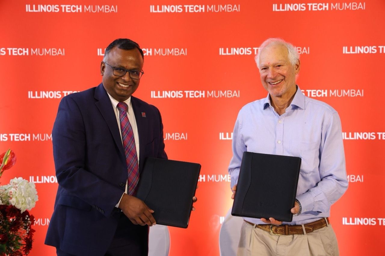 Two men smile for a photo holding portfolios containing a memorandum of understanding. They are in front of a backdrop with the words "Illinois Tech Mumbai" repeated.