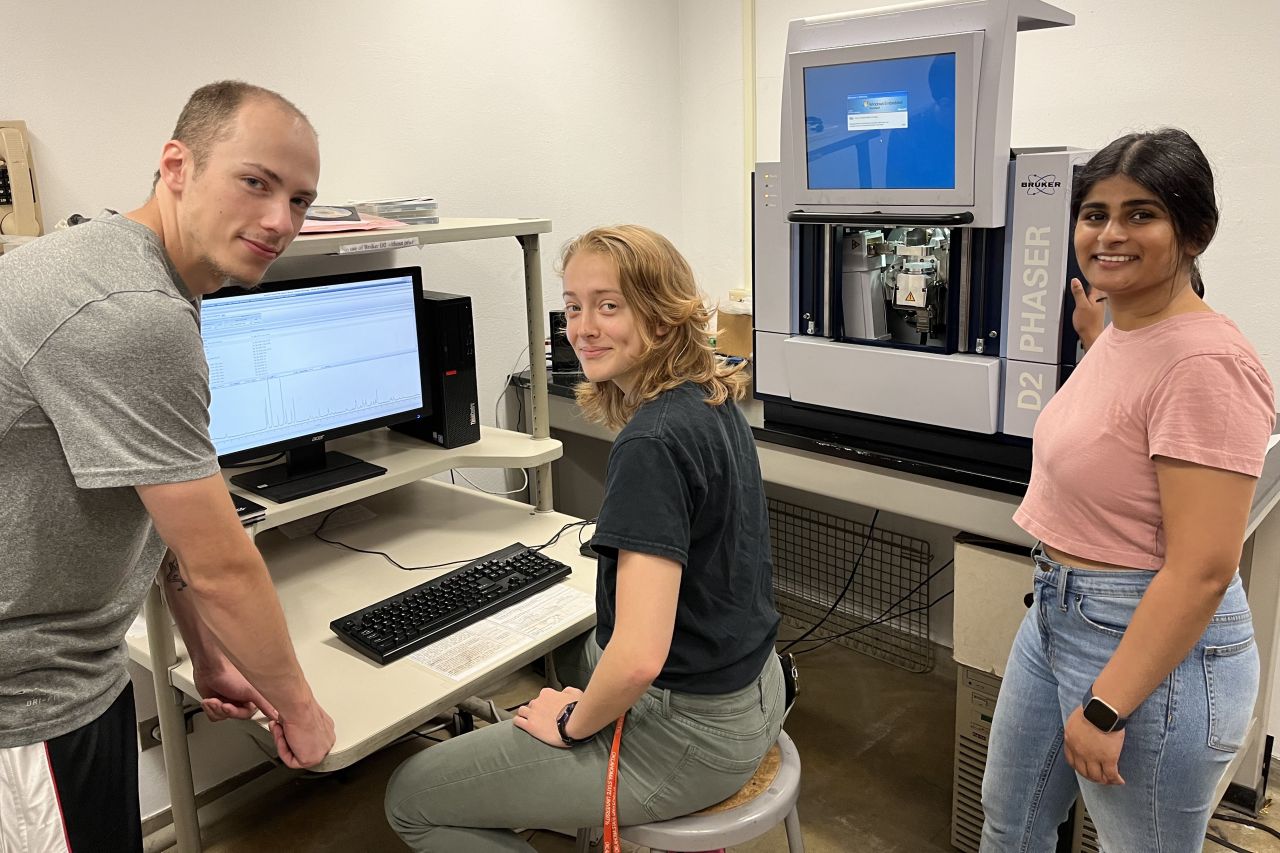 Three students pose for a photograph in front of a computer and a piece of lab equipment.