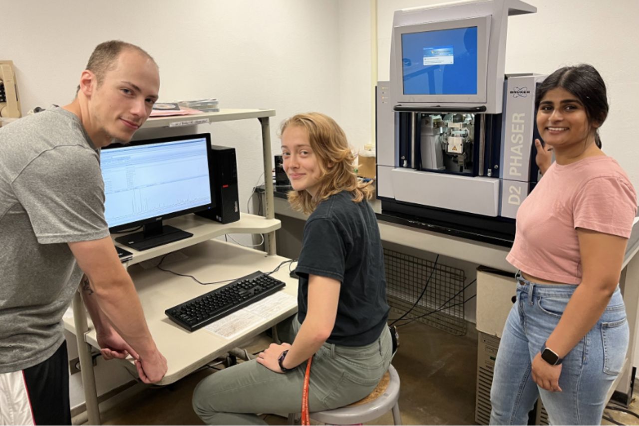 Three students pose for a photo in front of a computer and some lab equipment.