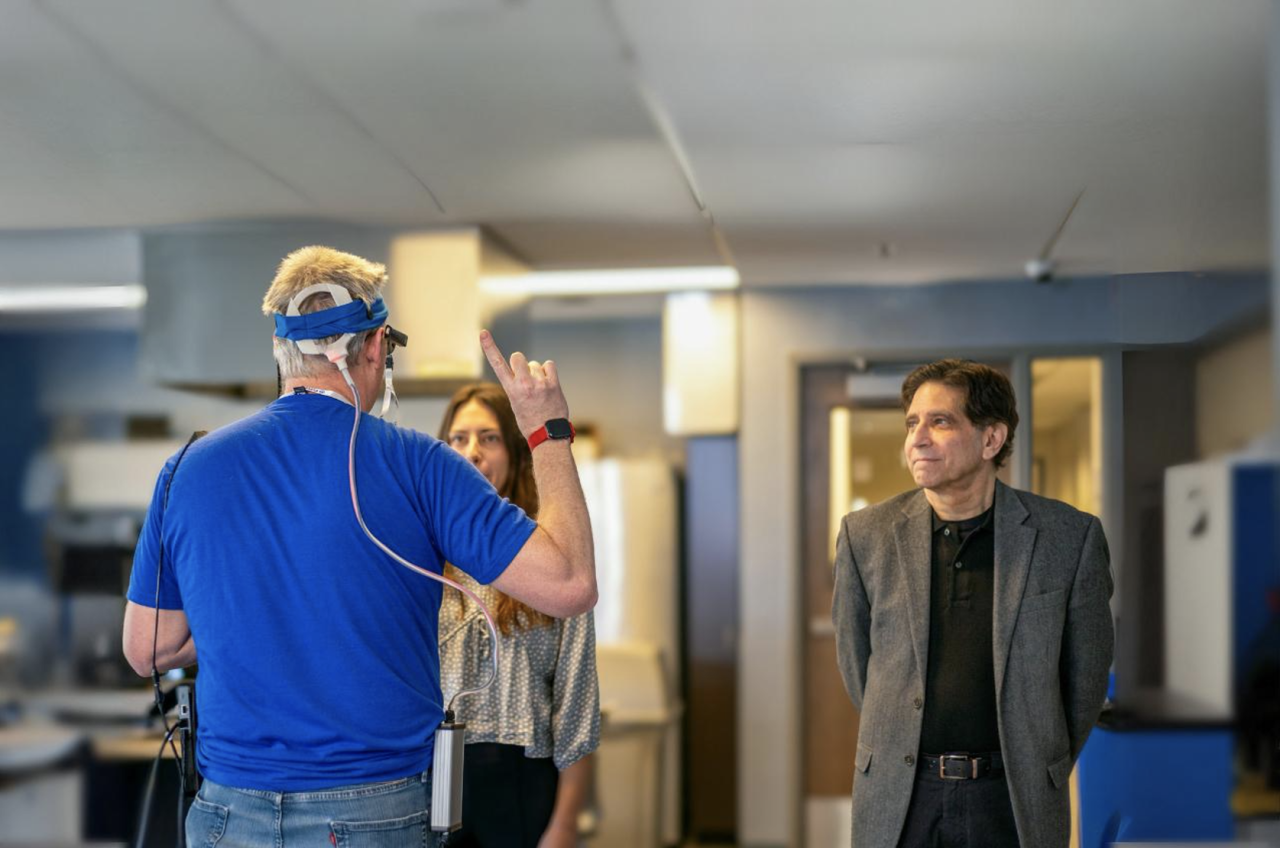 A man in a blue shirt wearing a device on his head holds up a finger in the air while facing away from camera and toward a woman who's partially obscured from the camera. To the right, a man in a black T-shirt and blazer looks at the man wearing the device. They are indoors and the background is in soft focus.