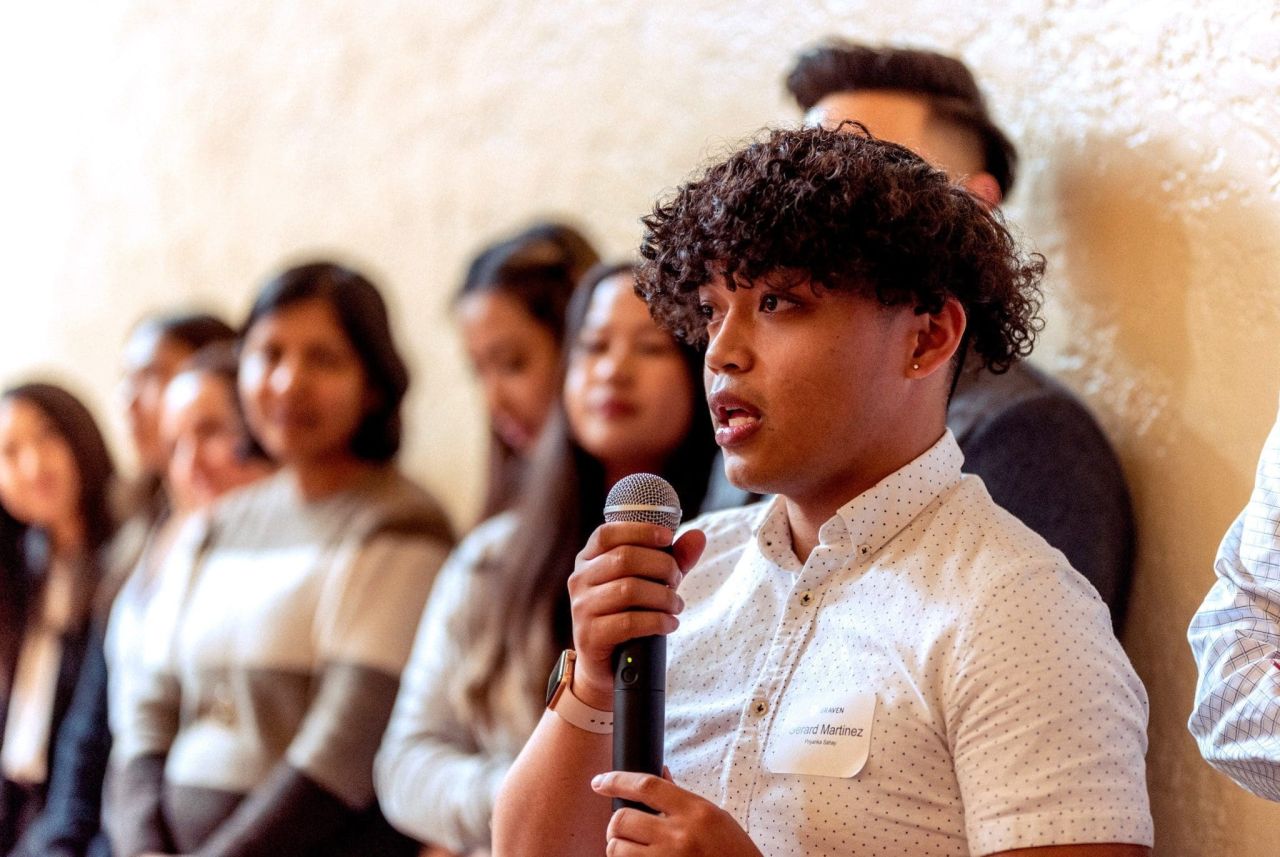 A young man in a white button-down shirt speaks into a microphone during an event as a group of attendees listens in the background.
