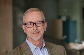 A man wearing dark glasses, a blue shirt and tweed shirt smiles in a portrait.