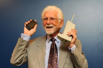 A man holds a large early cellphone in his left hand and a smaller, more modern cellphone in his right hand.