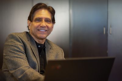 A man in a tweed jacket and dark shirt smiles in a portrait. 
