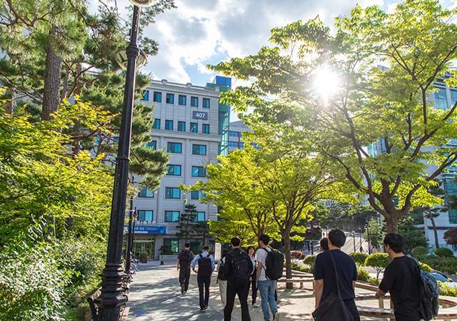 Sunlight filters through green trees as students walk toward a university building on the Hanyang University campus.