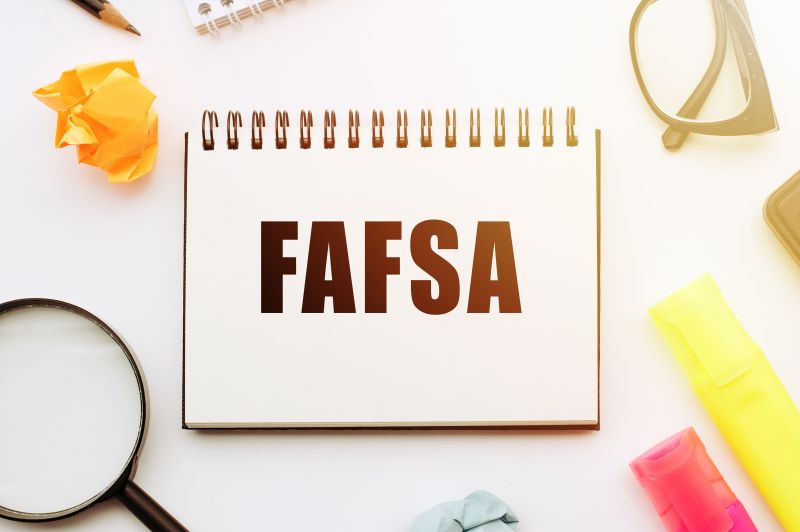 Notebook with the word FAFSA written surrounded by a magnifying glass, crumpled paper, glasses, and markers